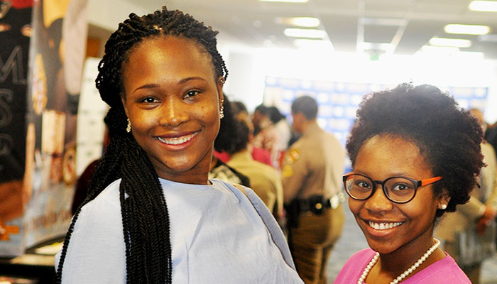 A tall Black young woman wearing long black braids with a white shirt stands next to a short Black young woman wearing black glasses, an afro, pearl earrings, pearl necklace, and pink v-neck sweater